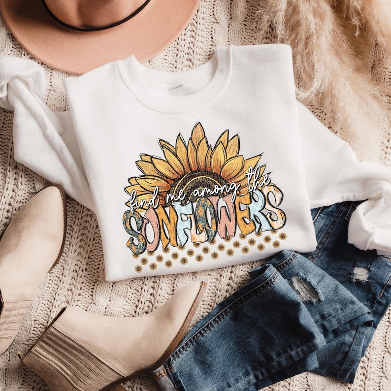 May include: White sweatshirt with a sunflower graphic and the text "find me among the SUNFLOWERS". The design features a large sunflower with yellow, orange, and brown petals. The sweatshirt is paired with blue jeans and beige boots.