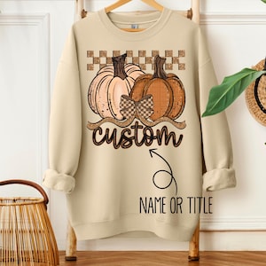 May include: A beige sweatshirt featuring a fall-themed design. The design includes two pumpkins, a bow, and the word "custom" in a script font. Below the word "custom" is an arrow pointing to the words "NAME OR TITLE".