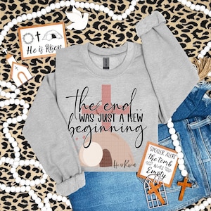 May include: A gray sweatshirt with the text "The end... was just a new beginning" and a graphic of a tomb with a cross. The text "He is Risen" is also included on the graphic.