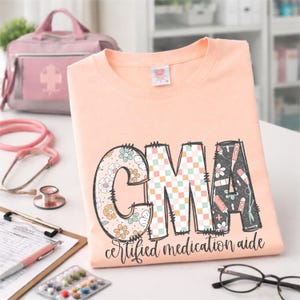 Puede incluir: Una camiseta color melocotón con las letras "CMA" en un diseño decorativo. La "C" está llena de motivos florales, la "M" con un patrón de cuadros y la "A" con herramientas médicas. El texto "certified medication aide" está debajo.