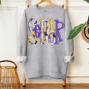 May include: A heather gray sweatshirt with the word "Senior" in large, colorful letters. The letters are in purple, yellow, and white, with a patterned design. The sweatshirt has long sleeves and a crew neck.