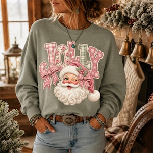 May include: A sage green long-sleeve sweatshirt featuring the word JOLLY in large, patterned block letters. Below the text is a graphic of a winking Santa Claus wearing a pink hat with a bow, accented by a pink bow on the left side of the text.
