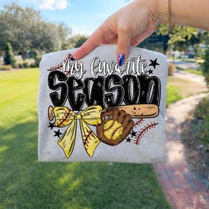 May include: A gray t-shirt featuring the text "my favorite SEASON" in a playful font. The design includes a yellow bow, a baseball glove, a baseball, and a baseball bat. Black stars and red stitching accents complete the design.