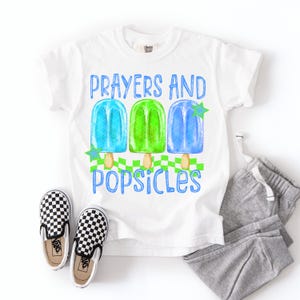 Puede incluir: Camiseta blanca con el texto azul "Prayers and Popsicles". La camiseta presenta un gr&aacute;fico de tres paletas de colores: azul, verde y azul. Un par de zapatos sin cordones a cuadros blancos y negros y un pantal&oacute;n de ch&aacute;ndal gris tambi&eacute;n est&aacute;n en la imagen.
