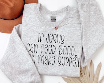 May include: A light gray sweatshirt with the text "If Jesus Can Feed 5000, I Make Supper" in black lettering. A brown hat and gold necklace are also visible.