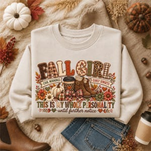 May include: A beige crewneck sweatshirt featuring a graphic print with the text FALL GIRL, COOL MORNINGS, EARLY SUNSETS, and THIS IS MY WHOLE PERSONALITY UNTIL FURTHER NOTICE. The design includes a coffee cup, boots, pumpkins, and flowers.