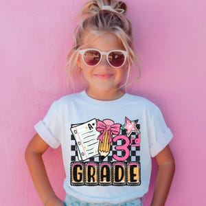 May include: A white t-shirt featuring a colorful graphic design for 3rd grade. The design includes a notebook, pencil, bow, star, and the words "3rd Grade" in a playful font. The shirt is worn by a young girl wearing sunglasses.