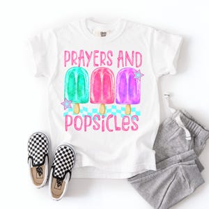Może przedstawiać: Biała koszulka z napisem "Prayers and Popsicles" i grafiką trzech kolorowych lod&oacute;w na patyku. Lody na patyku są zielone, r&oacute;żowe i fioletowe. Na zdjęciu znajdują się r&oacute;wnież czarno-białe szachownicowe buty wsuwane i szare spodnie dresowe.