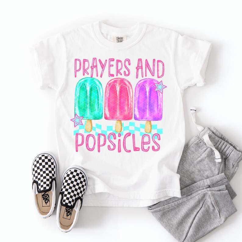 Może przedstawiać: Biała koszulka z napisem "Prayers and Popsicles" i grafiką trzech kolorowych lod&oacute;w na patyku. Lody na patyku są zielone, r&oacute;żowe i fioletowe. Na zdjęciu znajdują się r&oacute;wnież czarno-białe szachownicowe buty wsuwane i szare spodnie dresowe.