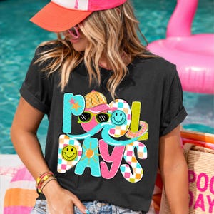 May include: Black t-shirt with the words "POOL DAYS" in colorful, playful letters. The design includes a cartoon character with sunglasses, a hat, and a mustache. The shirt is worn by a person.