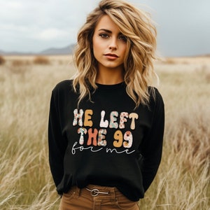 May include: A black long-sleeved sweatshirt with a colorful graphic that reads "He left the 99 for me". The model is wearing brown pants and has long blonde hair.
