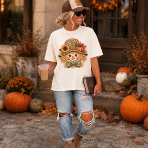 May include: An off-white short-sleeved t-shirt featuring a graphic of a scarecrow wearing a hat decorated with a sunflower, a pumpkin, and autumn leaves, paired with light blue distressed denim jeans with rips at the knees.