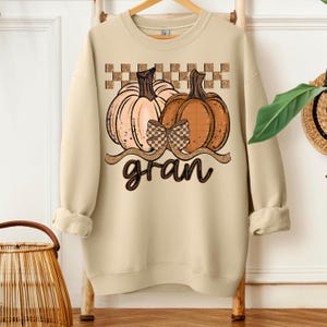 Peut inclure: Sweat-shirt beige avec un motif de deux citrouilles, un nœud à carreaux et le mot "gran". Les citrouilles sont orange et pêche, avec un motif à carreaux au-dessus. Thème automnal.