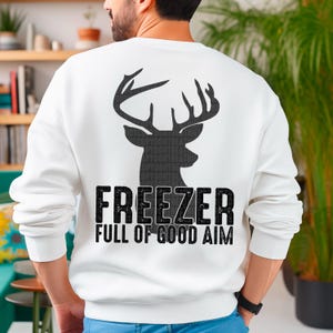 May include: White sweatshirt with a black deer head graphic and the text "FREEZER FULL OF GOOD AIM." The design features a silhouette of a deer's antlers and head. The sweatshirt is a casual, comfortable garment.