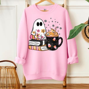 May include: Pink sweatshirt featuring a ghost wearing a floral sheet, stacked books, and a black mug with a pumpkin spice latte design. The ghost has black oval eyes and the mug has candy corn accents. The design is perfect for Halloween or fall.