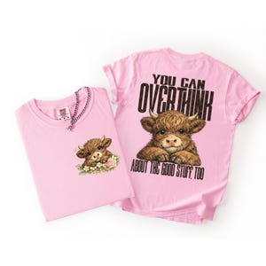May include: Two pink t-shirts. One shirt features a brown Highland cow graphic with flowers. The other shirt has a similar cow graphic and the text "YOU CAN OVERTHINK ABOUT THE GOOD STUFF, TOO."