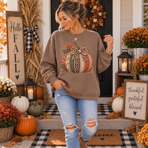 May include: A tan long-sleeve sweatshirt featuring a large graphic of a pumpkin decorated with various patterns including leopard print, plaid, and floral designs, accented with a central bow and autumn leaves, paired with light blue distressed jeans.