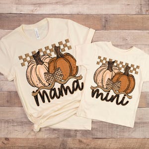 May include: Two cream-colored t-shirts with pumpkin graphics. One shirt says "Mama" and the other says "mini" in a cursive font. The pumpkins are brown and peach with a checkered pattern above them.