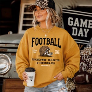 May include: A mustard yellow crewneck sweatshirt featuring a black graphic of a football helmet and the text FOOTBALL, LOVE BIG, PRAY HARD, TOUCHDOWNS, TAILGATES, & TRUSTING GOD, PROVERBS 3:5. The sweatshirt is paired with a white cup with GAME DAY text.