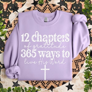 May include: A lavender sweatshirt with the text "12 chapters of gratitude 365 ways to live His Word" and a white cross.
