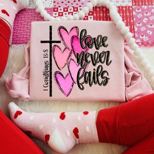 K&ouml;nnte beinhalten: Ein hellrosa Sweatshirt mit dem Text "love never fails" und dem Bibelvers "1 Korinther 13,8". Das Design umfasst ein schwarzes Kreuz und drei rosa Herzen mit goldenen Umrissen. Das Sweatshirt ist gefaltet.