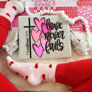K&ouml;nnte beinhalten: Ein hellgraues Sweatshirt mit dem Text "love never fails", einem Kreuz und rosa Herzen. Das Sweatshirt wird mit einer roten Hose und Socken mit roten Herzmustern getragen. Eine wei&szlig;e Pompon-Girlande befindet sich im Hintergrund.