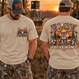 May include: A beige t-shirt featuring a small graphic on the front left chest and a large graphic on the back. Both graphics depict a deer head, pine trees, and a sunset with the text BUCK FEVER. The back includes the text THE SEASON WE WAIT FOR ALL YEAR.