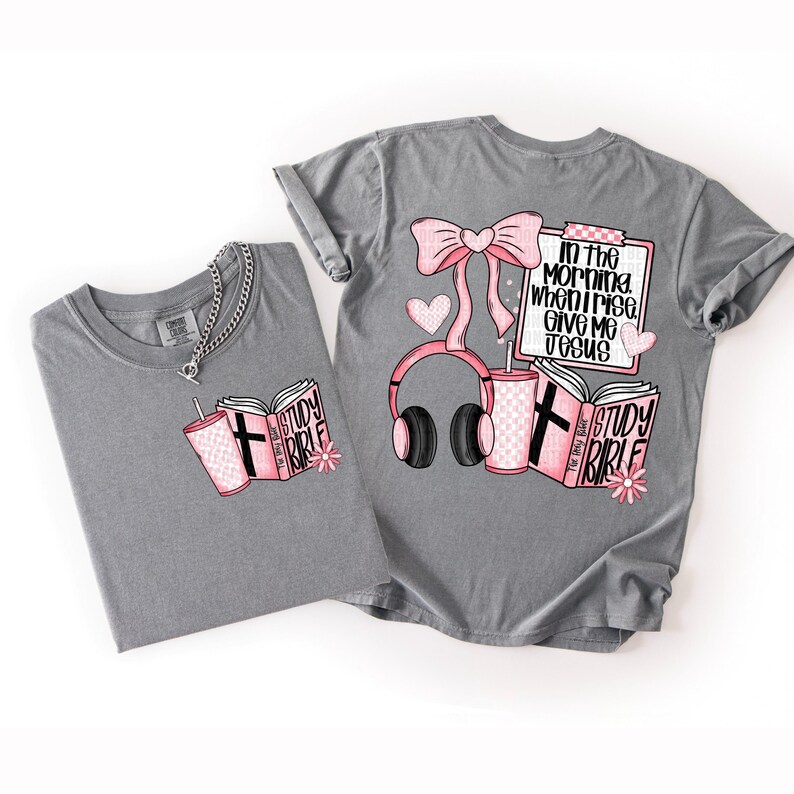 May include: Two gray t-shirts with pink and black graphics. One shirt features a book, a drink, and the words "Study Bible." The other shirt has a bow, headphones, a drink, a notepad with text, and a book. The text on the notepad says "In the morning when I rise, give me Jesus."