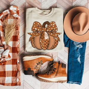 Puede incluir: Una sudadera crema con un diseño de calabaza y un lazo grande. La calabaza es marrón con lunares y un patrón de cuadros. La imagen también incluye una camisa a cuadros, botas marrones, un sombrero y vaqueros azules.