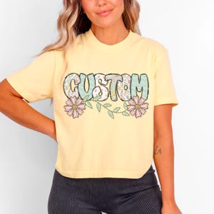 Op de afbeelding: Een geel gecropped t-shirt met een bloemenpatroon waarop in een retro-lettertype "CUSTOM" staat.