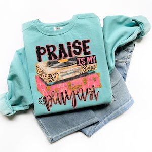 May include: A turquoise sweatshirt with the text "PRAISE IS MY PLAYLIST" in black and pink script. The graphic features a record player and stacked pink and leopard print suitcases. The sweatshirt is paired with denim shorts.