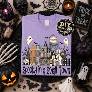 May include: A light purple t-shirt featuring a graphic of a small town with a water tower labeled Petersburg, a sign reading 47567, and the text Spooky in a Small Town. The design includes ghosts, bats, and various buildings in orange, purple, and yellow.