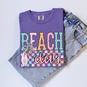 May include: A purple Comfort Colors t-shirt with a "Beach Day" graphic. The graphic includes a palm tree, surfboard, and a checkered pattern in pastel colors. The shirt is paired with light-wash denim jeans.