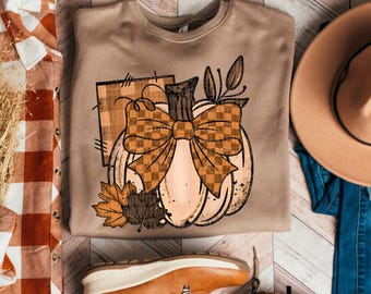 Puede incluir: Sudadera beige con un diseño de calabaza con un gran lazo a cuadros, hojas y un cuadrado a cuadros. La imagen incluye un sombrero, botas y una camisa a cuadros, todo con temática otoñal.