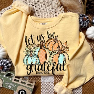 May include: A light yellow sweatshirt with the words "let us be grateful" and a graphic of pumpkins in shades of orange, teal, and gold. The text "Hebrew 12:28" is below the word "grateful". The sweatshirt has long sleeves.