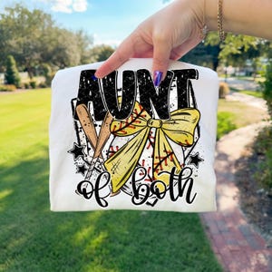 以下が含まれることがあります： 野球をテーマにしたグラフィックの白いTシャツ。デザインは、黒で「AUNT」という言葉、野球のバット、黄色のリボン、そして「of both」というフレーズを特徴としています。星と野球のステッチがデザインを完成させます。