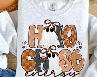 May include: A light grey sweatshirt featuring the phrase "Hi Ghost Energy". The text is in orange and brown with a checkered pattern. Two ghost figures with bows are also visible on the sweatshirt.