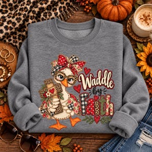 Puede incluir: Una sudadera gris jaspeado de cuello redondo con el gráfico de un pato con gafas y un lazo estampado. El pato sostiene una cruz decorada con flores. El texto Waddle by Faith aparece en letras con estampado de cuadros rojos y negros y leopardo.
