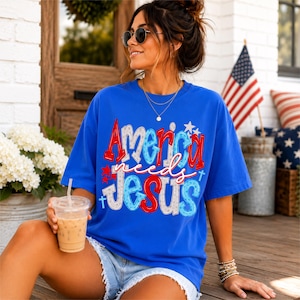 K&ouml;nnte beinhalten: Ein k&ouml;nigsblaues T-Shirt mit dem Text "America needs Jesus" in Rot, Wei&szlig; und Blau. Das Shirt hat einen Rundhalsausschnitt und kurze &Auml;rmel. Die Person tr&auml;gt eine Sonnenbrille und Jeansshorts.