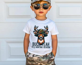May include: A young child wearing a white t-shirt with a deer graphic and the text "BUCK YEAH IT'S HUNTING SEASON." The child is also wearing camouflage shorts, a camouflage cap, and sunglasses.