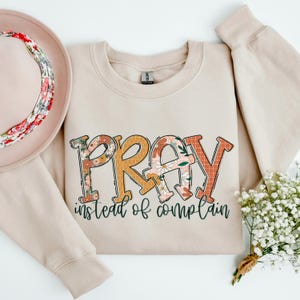 May include: A beige sweatshirt with a floral print design that says "Pray instead of complain".