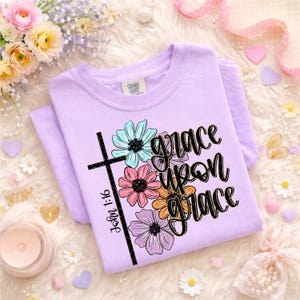 May include: A light purple t-shirt with the words "grace upon grace" in black script, accented by a black cross and colorful floral graphics. The text "John 1:16" is printed vertically on the cross. The shirt is folded.