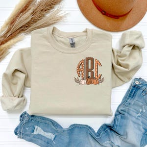 May include: A beige sweatshirt featuring a monogram design with the letters "ABC" in a fall-themed pattern. The design includes pumpkins and leaves. The sweatshirt is paired with blue jeans and a brown hat.