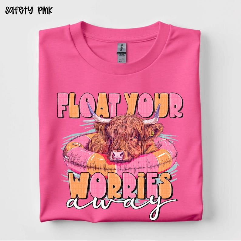 May include: Safety pink t-shirt with a graphic of a Highland cow in a pink and orange inner tube. The text "FLOAT YOUR WORRIES AWAY" is above and below the cow. The shirt is folded on a white surface.