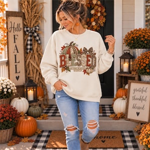 May include: A cream-colored crewneck sweatshirt featuring a graphic print of pumpkins, autumn leaves, and a bow. The text reads BLESSED BEYOND MEASURE. The sweatshirt is paired with light blue distressed denim jeans with rips at the knees.