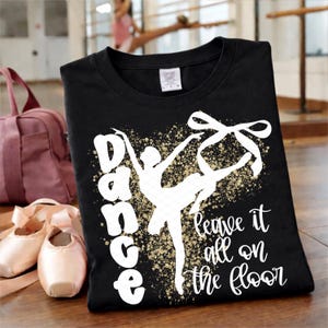 Könnte beinhalten: Schwarzes T-Shirt mit einer weißen Grafik einer Ballerina und den Worten "Dance leave it all on the floor". Eine rosa Sporttasche und Ballettschuhe liegen neben dem Shirt.