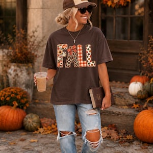 May include: A dark brown short-sleeve t-shirt featuring the word FALL in large block letters. Each letter has a different pattern including sunflowers, leopard print, red plaid, and polka dots. The shirt is paired with light blue distressed jeans.