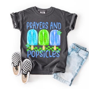 Puede incluir: Camiseta gris oscuro con las palabras "Prayers and Popsicles" y un gr&aacute;fico de tres polos de colores. La camiseta se combina con unos pantalones cortos vaqueros azul claro y unos zapatos sin cordones a cuadros blancos y negros.