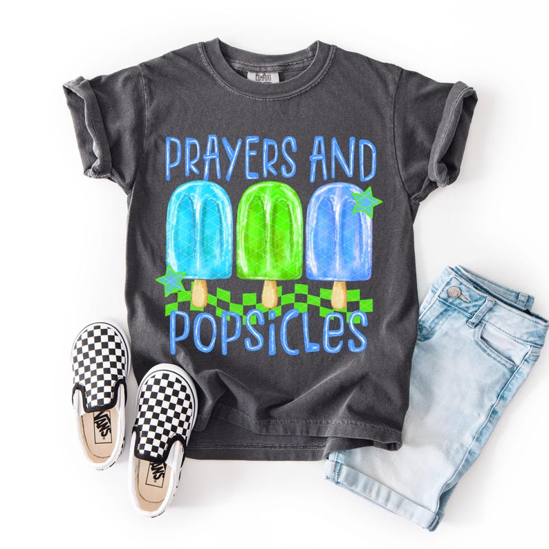 Puede incluir: Camiseta gris oscuro con las palabras "Prayers and Popsicles" y un gr&aacute;fico de tres polos de colores. La camiseta se combina con unos pantalones cortos vaqueros azul claro y unos zapatos sin cordones a cuadros blancos y negros.