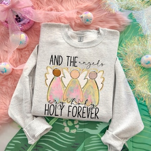 Puede incluir: Una sudadera gris claro con el texto "AND THE angels cry WHOA holy FOREVER" y un gráfico de tres ángeles con halos y alas doradas. Los ángeles tienen colores rosa, amarillo y marrón. La sudadera está sobre una superficie rosa y esponjosa.